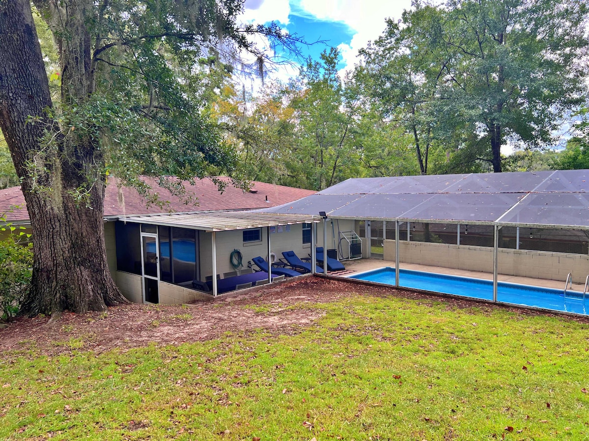 A spacious backyard view features a screened-in pool area with a blue pool, surrounded by green grass and trees. The home is visible with a covered patio hosting lounge chairs, offering a serene outdoor space for relaxation.
