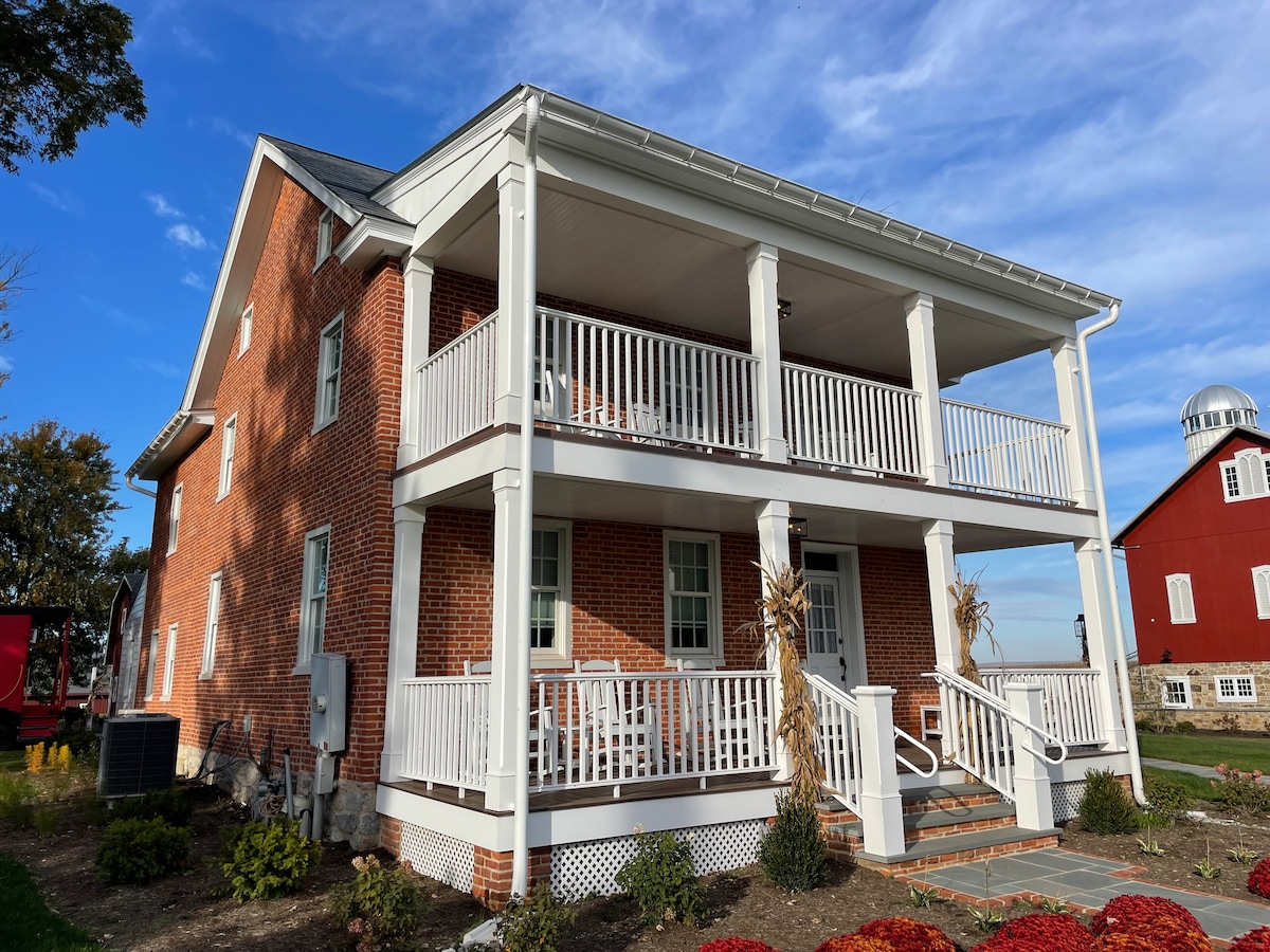 The exterior view of the farmhouse showcases red brick walls and two covered porches with white railings. Steps lead up to the entrances, surrounded by well-maintained landscaping and seasonal flowers. A classic red barn is visible in the background against a clear blue sky.