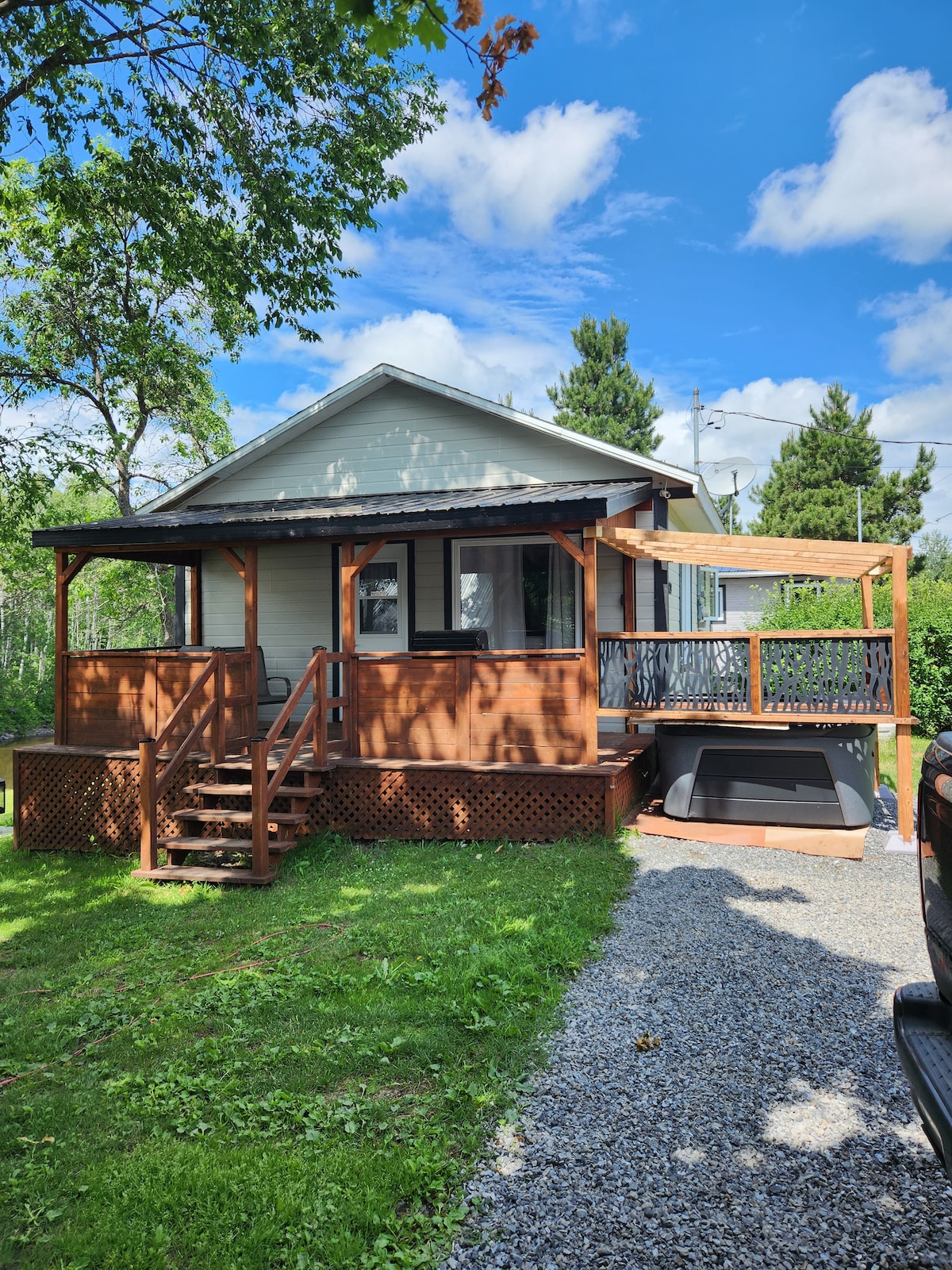 A charming chalet is set against a backdrop of blue skies, featuring a welcoming wooden porch. Steps lead to the entrance, and a hot tub is positioned on the side. Lush green grass surrounds the cabin, enhancing the serene outdoor environment.