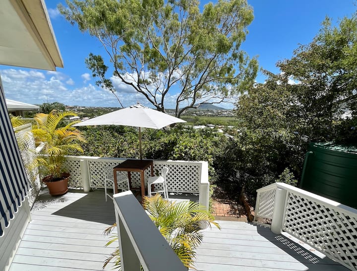 The image shows a bright and sunny outdoor deck area with light gray wooden flooring. A small table with two chairs is shaded by a white umbrella, surrounded by potted plants and lush greenery. The view overlooks a distant landscape of rolling hills and houses, with a blue sky filled with light clouds above. The deck features white lattice railings and leads down to a garden path, creating a peaceful, inviting spot to enjoy the scenery and the outdoors.