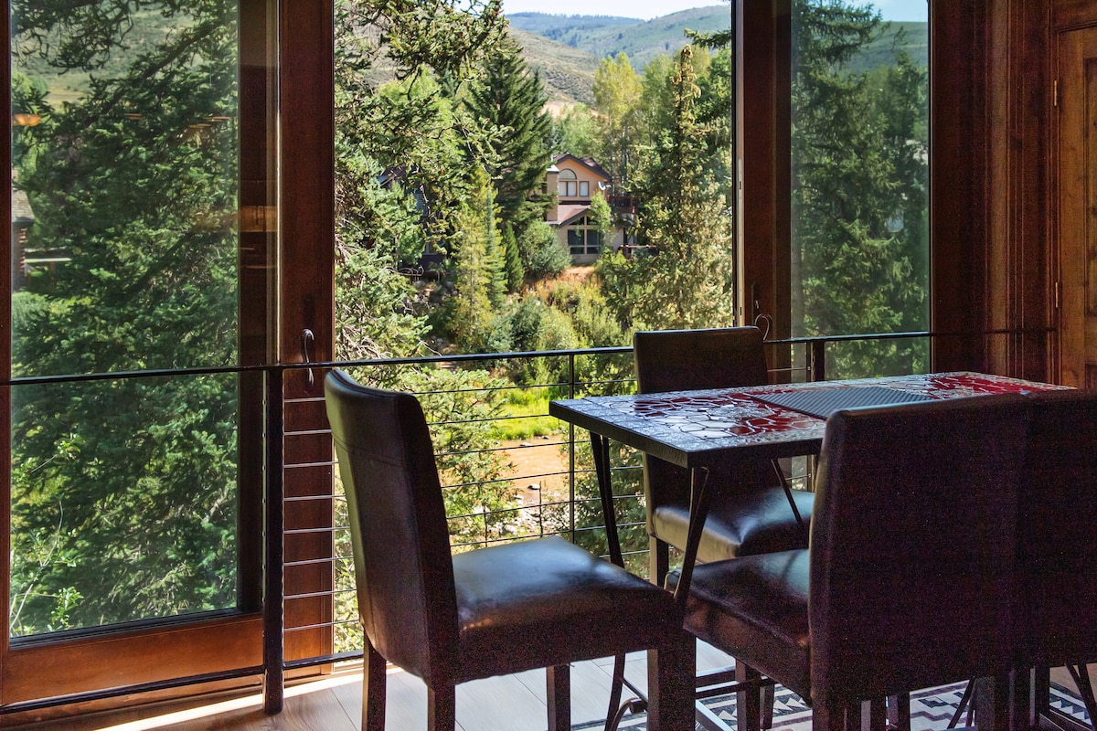 A dining area features a table surrounded by four dark leather chairs, overlooking expansive windows that frame a lush view of the surrounding trees and distant mountains. Natural light enhances the inviting space.