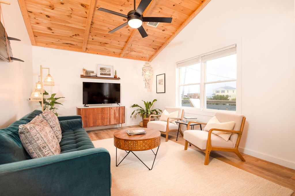 A spacious den is featured with a cozy, teal sofa and two light-colored armchairs. A round coffee table sits in the center, complemented by a wooden entertainment unit housing a flat-screen TV. Natural light streams in from two large windows, illuminating the wood-paneled ceiling.