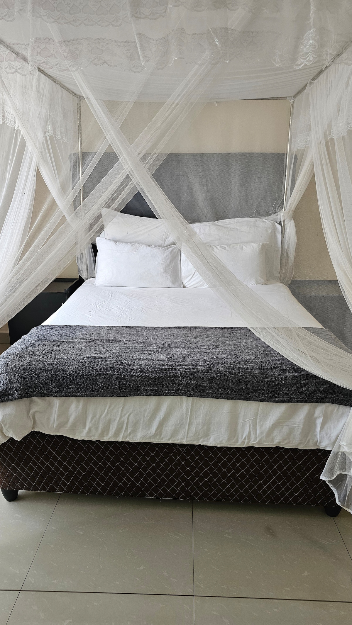 A spacious bed is draped in delicate white mosquito netting, creating a serene sleeping space. Soft, white pillows are arranged neatly against the headboard, which is complemented by a subtle grey backdrop. A light grey blanket is casually layered over the bed, adding a touch of comfort.