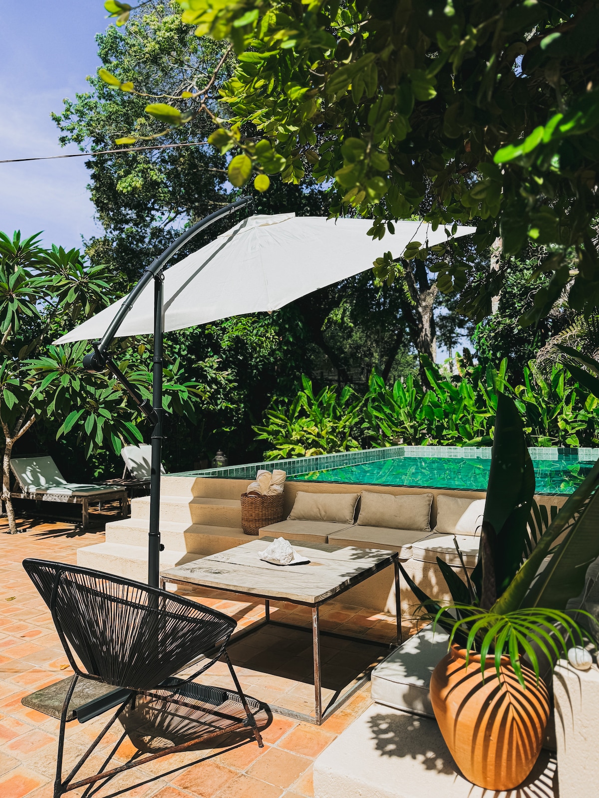 A shaded outdoor seating area is presented beside a clear pool, surrounded by lush greenery. A comfortable couch is positioned on a tiled patio, with a table in front and a chair nearby. A large umbrella provides relief from sun exposure.
