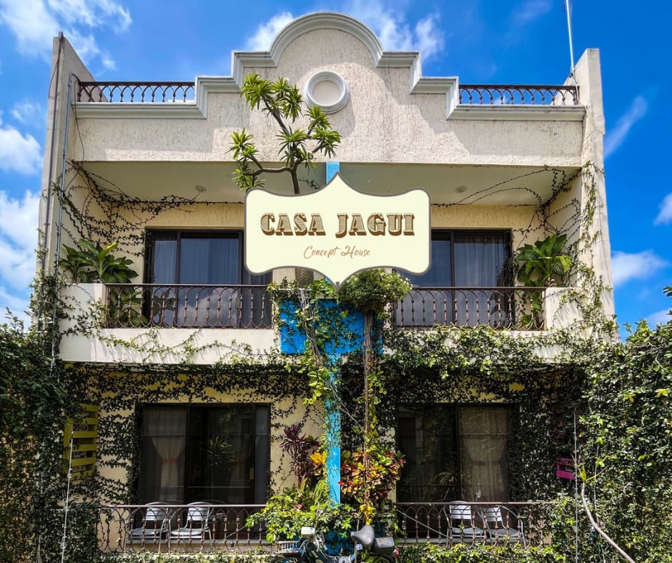 The exterior of Casa Jagui showcases a two-story building adorned with climbing greenery. Large windows allow natural light to flood the interior. A sign displays the name 'Casa Jagui,' welcoming guests. Outdoor seating is visible, offering a space for relaxation amongst the surrounding plants.