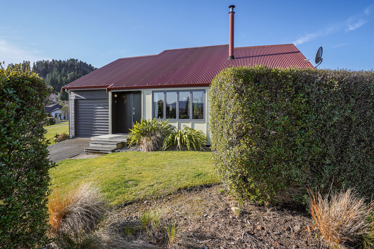 A single-story villa is showcased with a sloped, red metal roof and light green accents. The entrance is framed by manicured shrubbery, and steps lead up to a welcoming front door. A clear blue sky is visible above, enhancing the home's inviting façade.