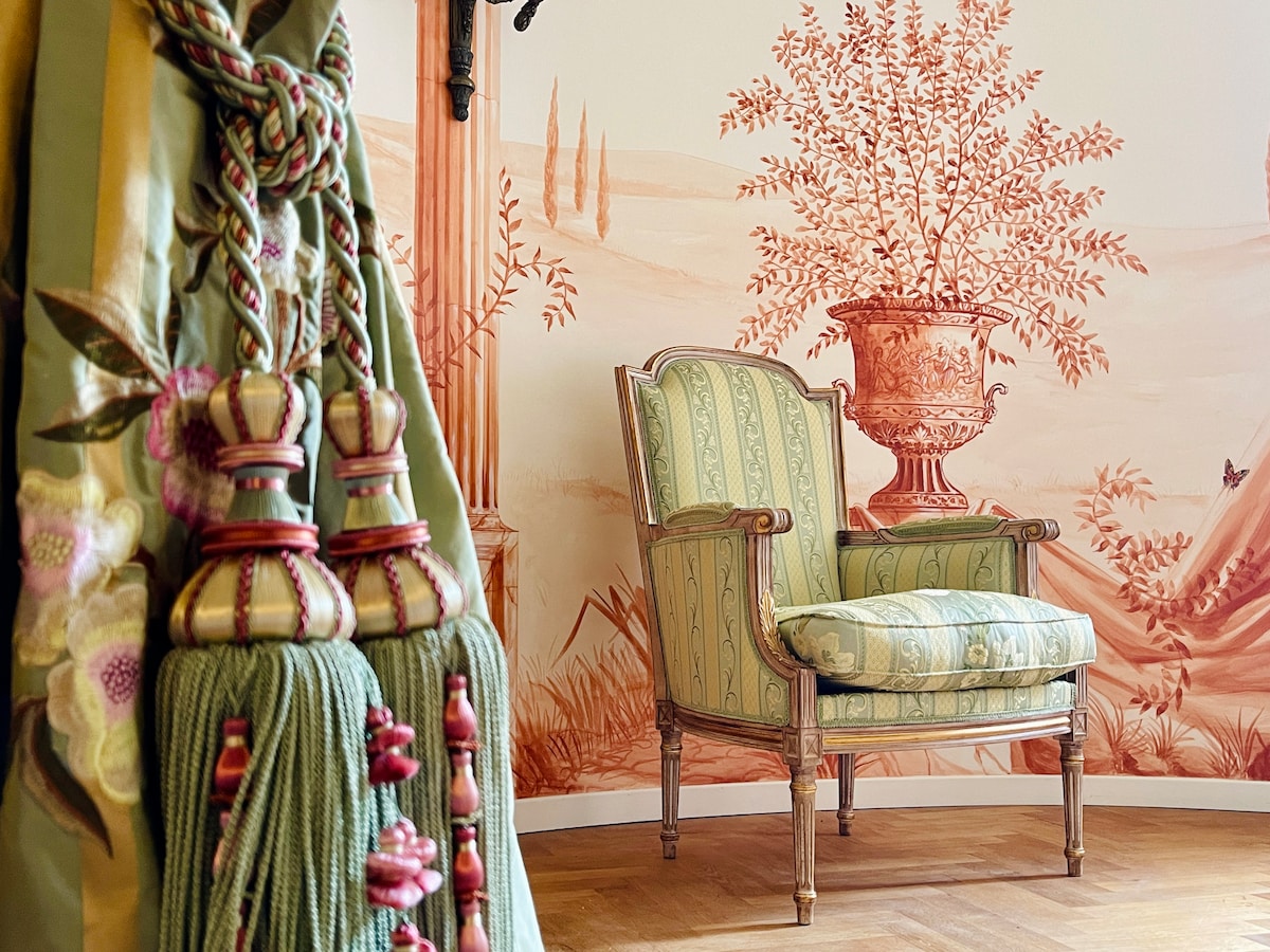 A charming armchair with a striped upholstery is positioned in front of a mural depicting a classical scene with foliage and a vase. Decorative tassels in muted tones hang nearby, adding a touch of elegance to the interior.