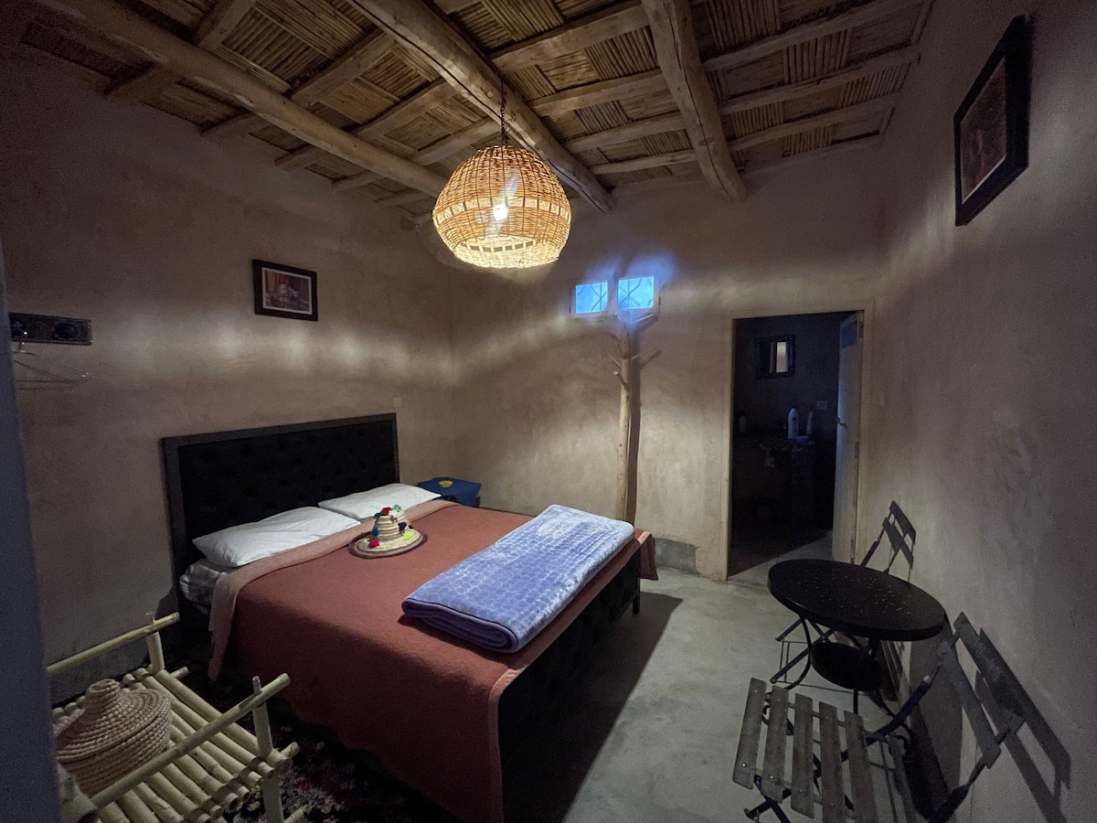 The double room features a spacious bed topped with a folded blanket. A round pendant light hangs from the ceiling, illuminating the earthy walls. Two chairs and a small table are arranged in one corner, while a door leads to a private bathroom.