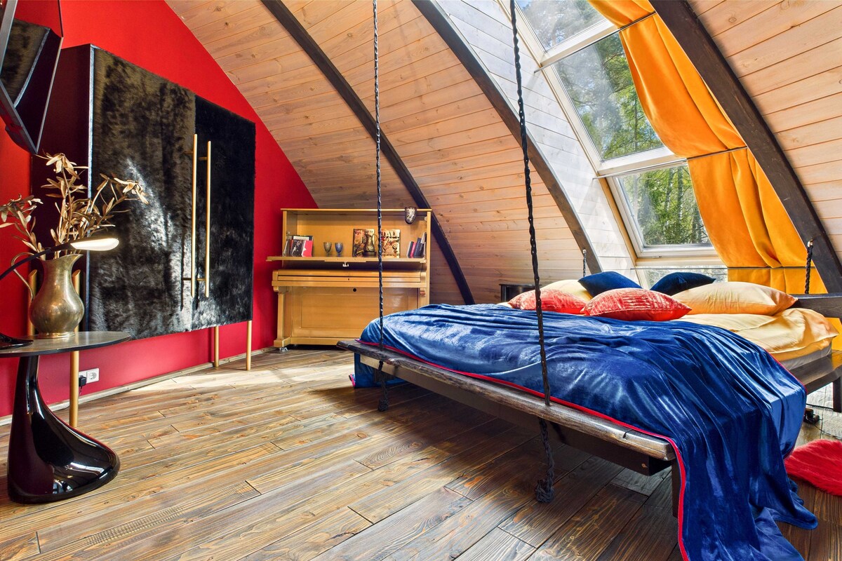 A vibrant room features a hanging bed draped with colorful pillows and a plush blanket. Large triangular windows offer natural light, showcasing wooden beams and flooring. A sleek black cabinet and a stylish desk enhance the artistic theme of the space.