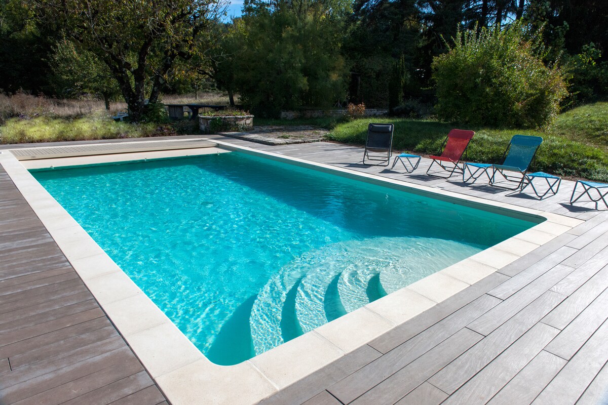 A heated outdoor pool is surrounded by a wooden deck and framed by lush greenery. Four lounge chairs in vibrant colors are arranged neatly along the edge of the pool, inviting relaxation. Sunlight reflects off the clear blue water, creating a serene atmosphere.
