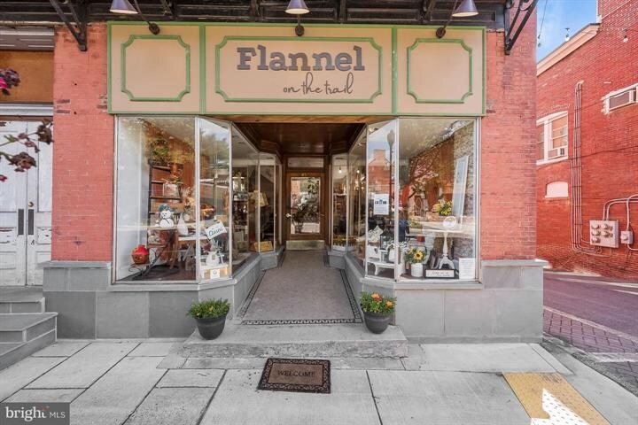 An inviting storefront presents large glass windows showcasing an assortment of home goods and decor. Potted plants are positioned at the entrance, adding a touch of greenery. The surrounding brick buildings contribute to the urban ambiance of the location.