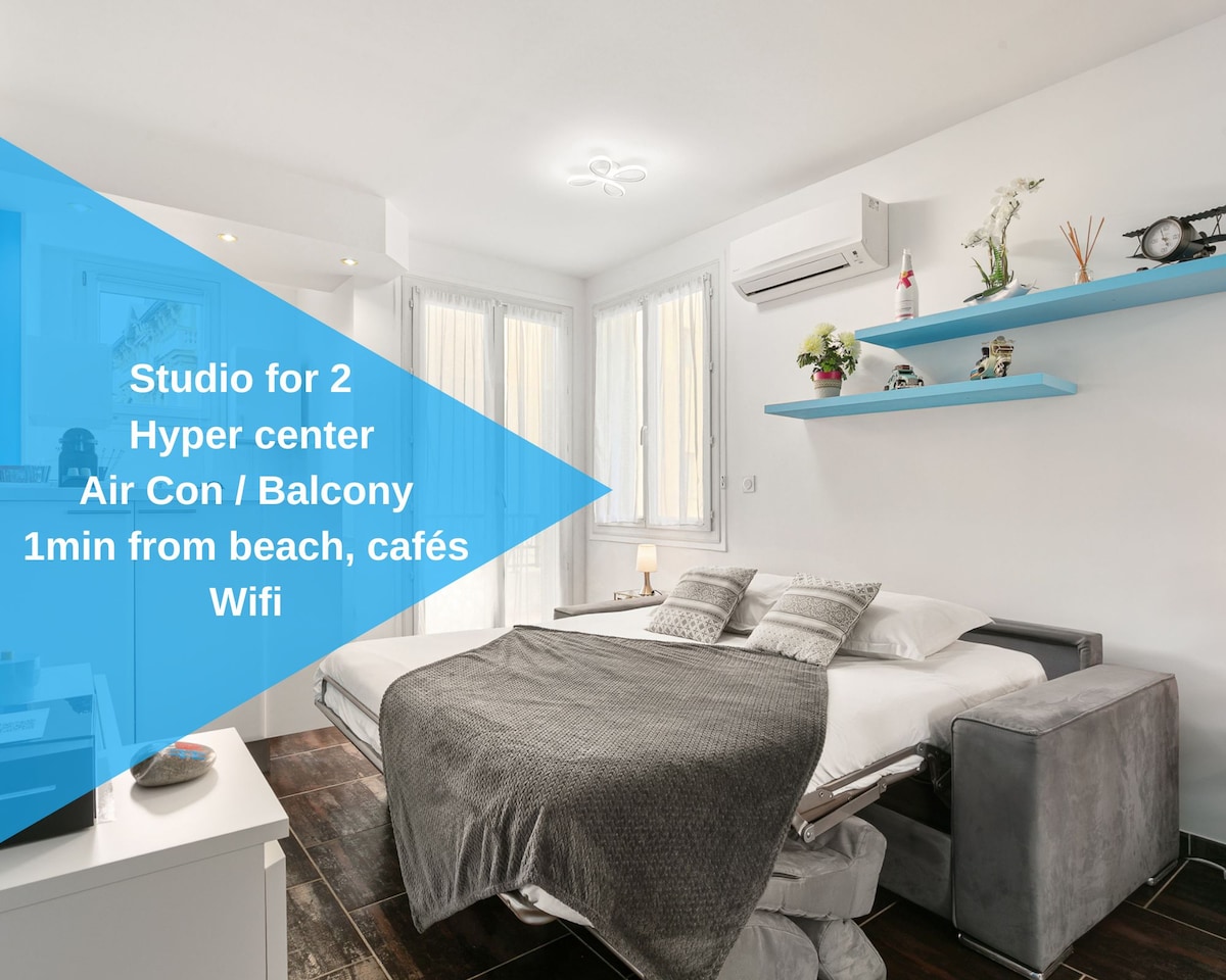 A bright studio space is designed for two, showcasing a spacious bed with a gray blanket. Natural light fills the room through large windows, and a small balcony provides outdoor access. Shelving holds decorative items, and air conditioning is visible for comfort.