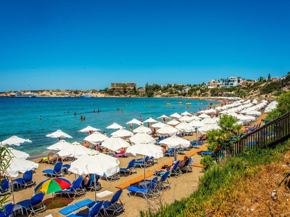 The view showcases a sandy beach lined with rows of white umbrellas and blue sun loungers. Crystal clear water gently laps against the shore, with beachgoers enjoying the sun and swimming. A variety of water sports activities can be observed in the distance, against a backdrop of a clear blue sky.