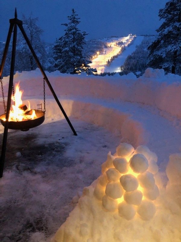 An inviting outdoor area is presented, featuring a fire pit surrounded by a low snow wall. A path of illuminated snow leads up the hillside, while a structure made of lighted snowballs adds a whimsical touch to the scene. Trees are visible in the background.