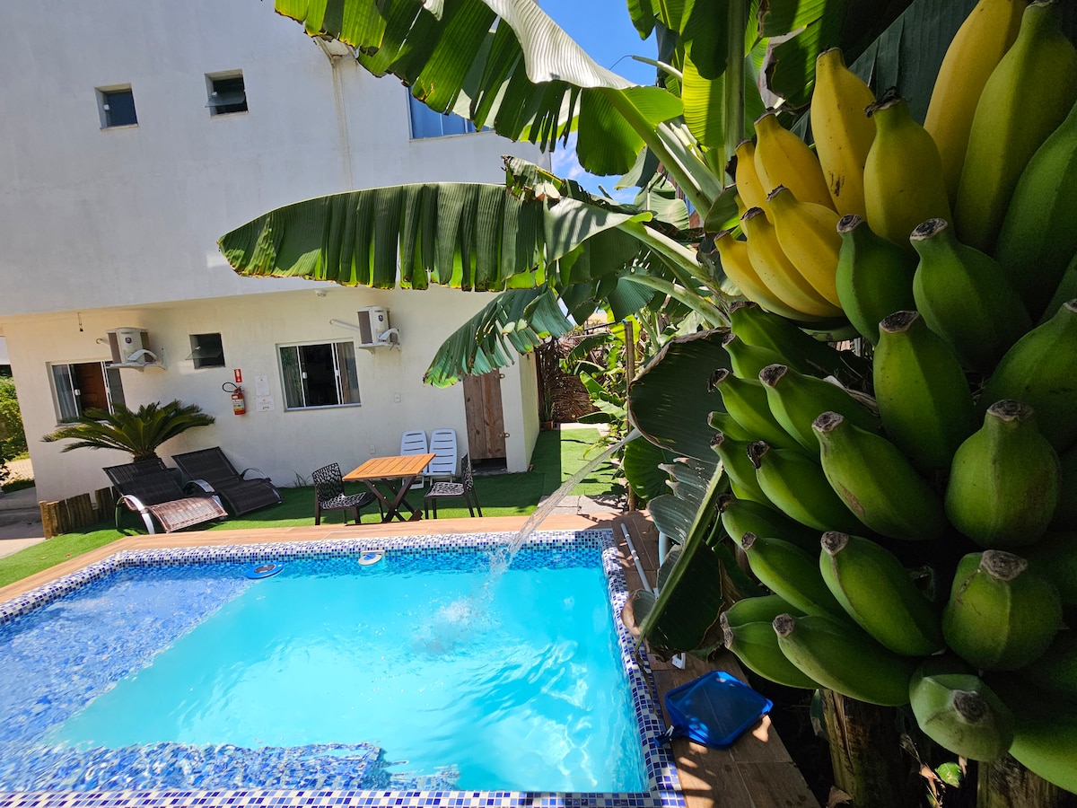 A refreshing pool features a water fountain surrounded by vibrant greenery. In the background, a spacious outdoor dining area and lounge chairs are visible. The property is complemented by clusters of bananas hanging from a nearby tree, enhancing the tropical ambiance.