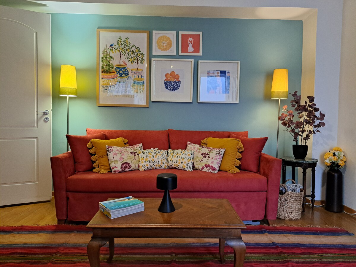 A vibrant seating area features a red sofa adorned with decorative cushions, complemented by a wooden coffee table. Colorful artwork hangs on the wall above, while soft lighting is provided by two standing lamps. A woven basket and an arrangement of flowers add warmth to the space.
