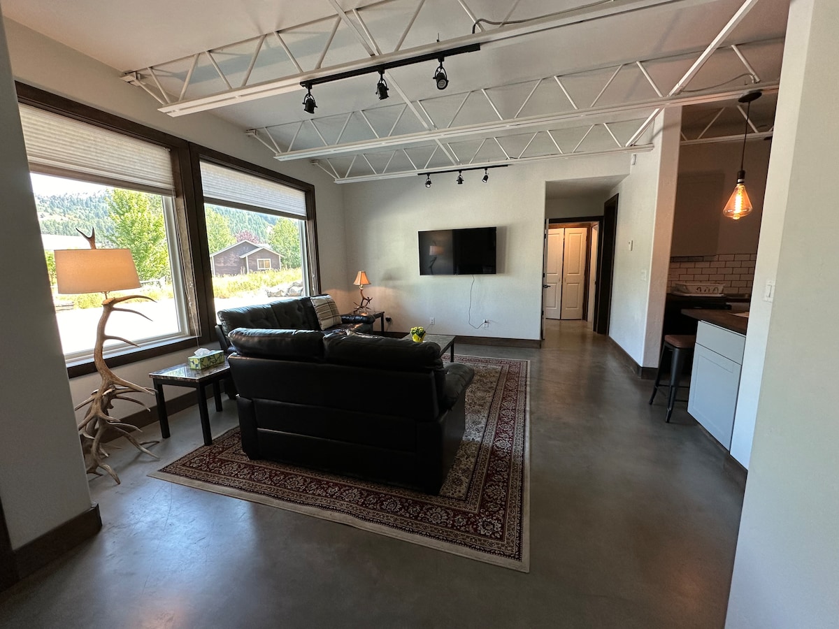 A spacious living area features large windows, allowing ample natural light and scenic views. Modern furniture is arranged with a leather couch and a small table. A flat-screen television is mounted on the wall, and a decorative rug lies on the polished concrete floor.