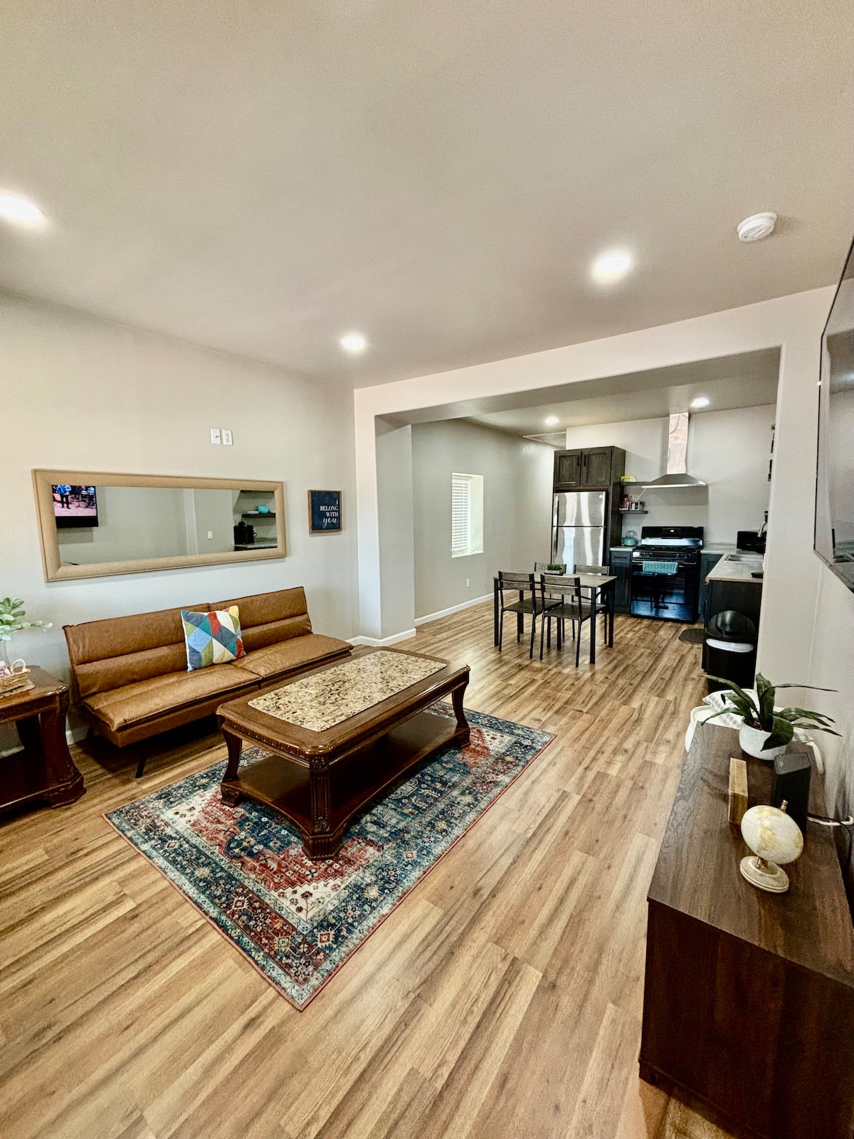 A living area displays a brown sofa paired with a coffee table featuring a patterned surface. A dining table with chairs occupies the space near an open kitchen, where modern appliances are visible. Natural light illuminates the wood flooring and neutral-colored walls.
