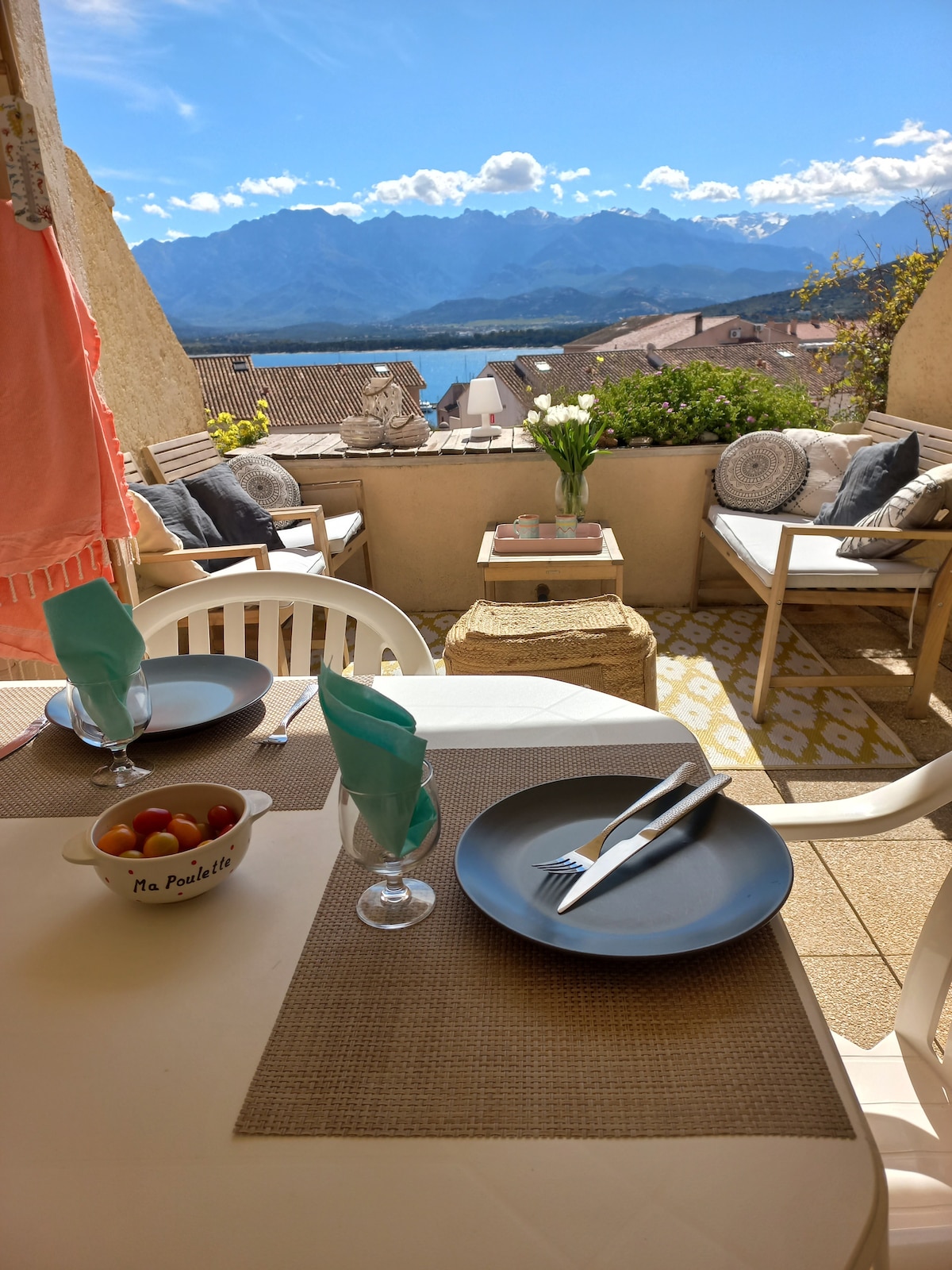 A terrace is presented, featuring a dining table set for two, with plates, silverware, and a bowl of fresh fruit. Comfortable seating areas are arranged to enjoy the mountain and bay views. Bright blue skies enhance the inviting atmosphere.