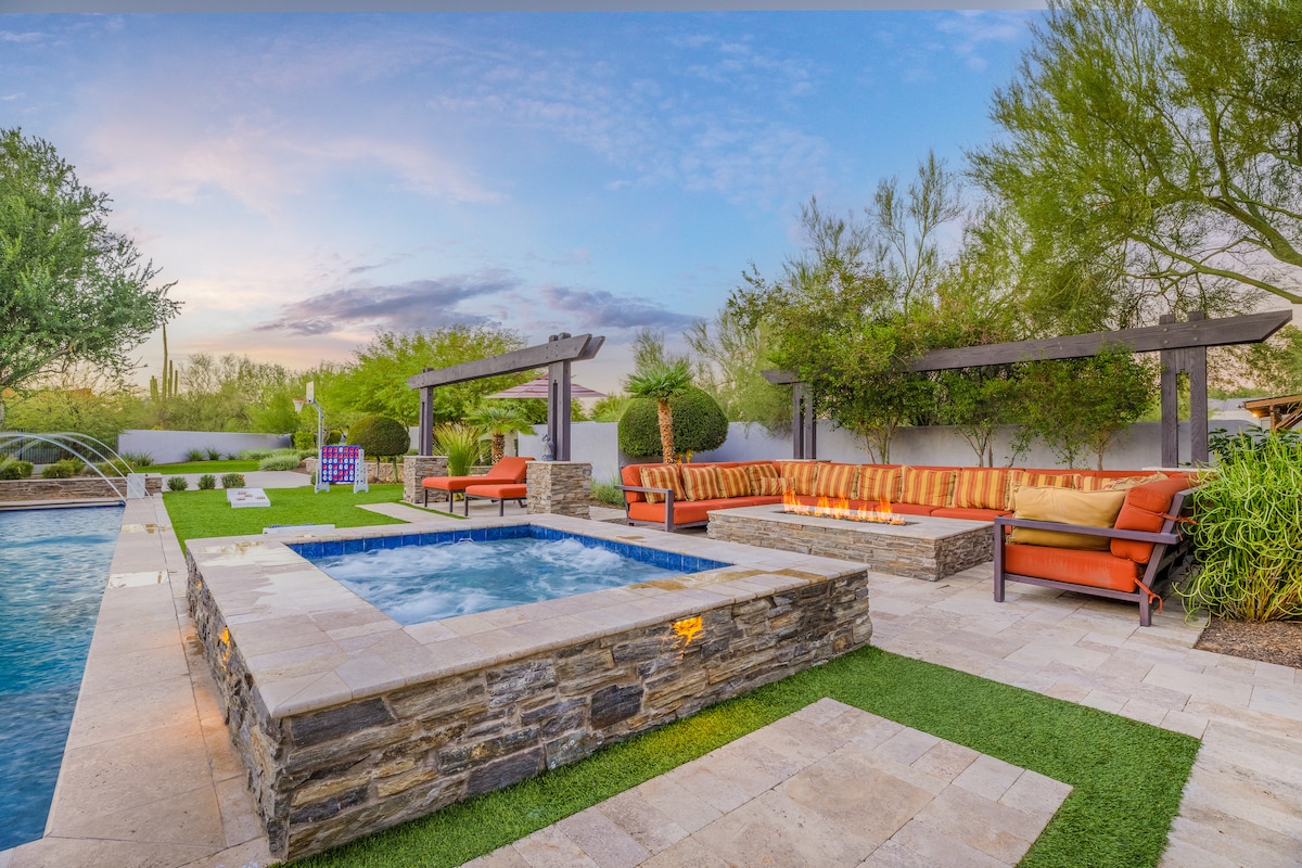 An outdoor area features a heated spa surrounded by stonework and a spacious seating area with plush sofas. Lush greenery and trees provide a natural ambiance, while the pool reflects the sky's hues. Pathways of grass and stone tiles create a seamless flow between elements.