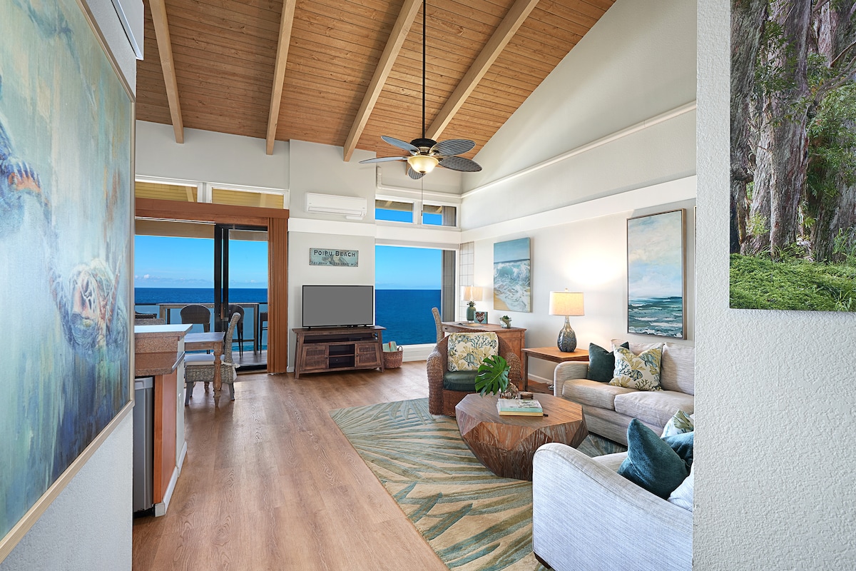 The living area features a high, wooden ceiling and an open layout, with large windows overlooking the ocean. A comfortable sofa is paired with a unique coffee table made of natural wood. Art pieces adorn the walls, enhancing the serene atmosphere.