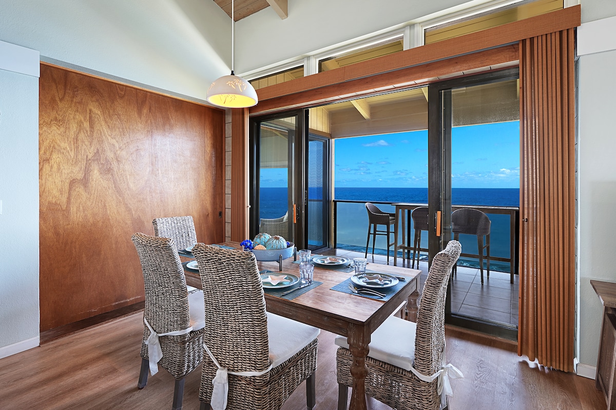 A dining area features a wooden table surrounded by four textured chairs, with place settings for dining. Floor-to-ceiling glass doors open to a lanai, allowing views of the ocean and horizon beyond, enhancing the indoor-outdoor experience.
