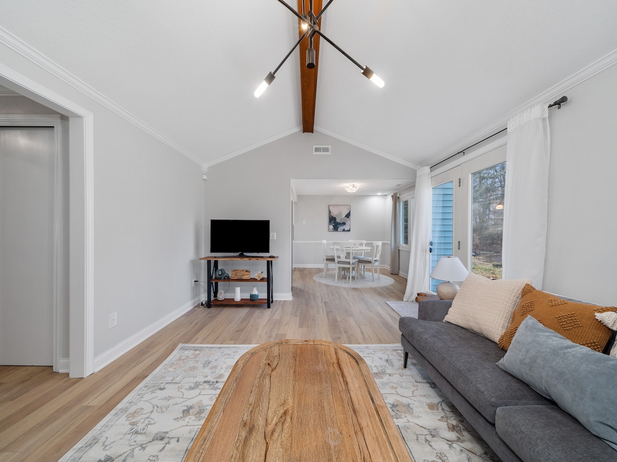 A well-lit living area features a spacious gray sofa adorned with neutral-toned cushions. A wooden coffee table sits in the center, while a television is mounted on the wall opposite. Large glass doors lead to the outdoor space, creating a connection to nature.