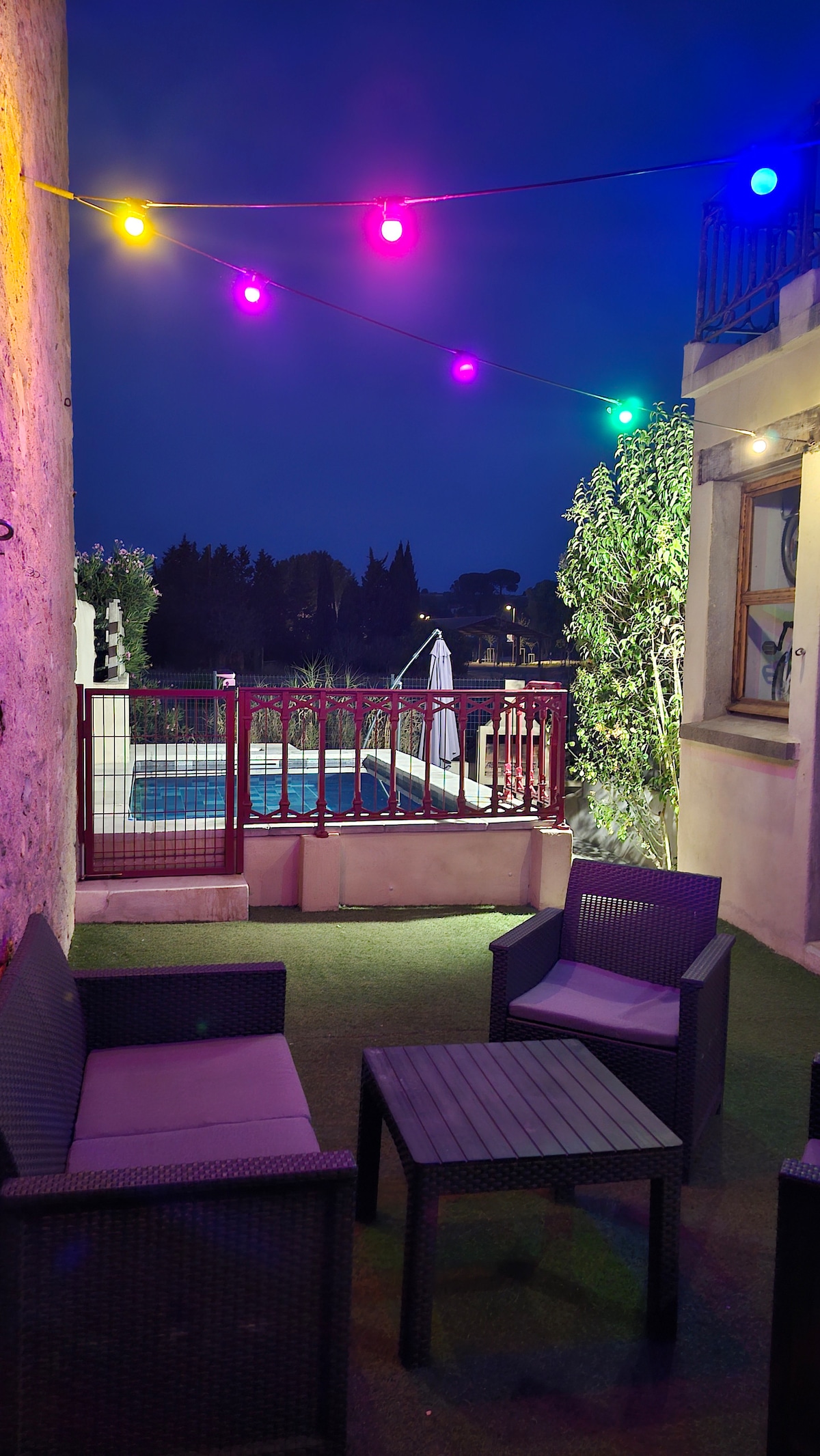 A cozy outdoor seating area is featured, illuminated by colorful string lights. The space includes a small table and four chairs, surrounded by greenery. A glimpse of a swimming pool can be seen in the background, contributing to a relaxed atmosphere.