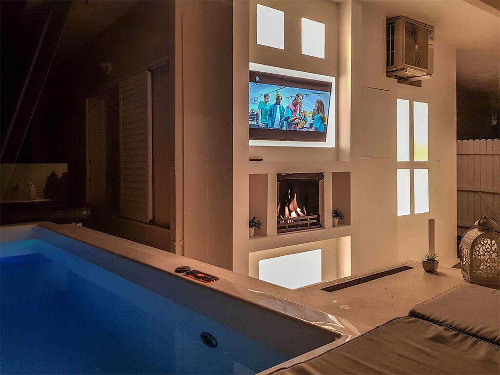 An indoor relaxation area is highlighted by a softly glowing indoor Jacuzzi and a modern fireplace. A mounted television displays a program, while large windows offer a glimpse of the exterior. A comfortable sunbed is positioned nearby for leisure.