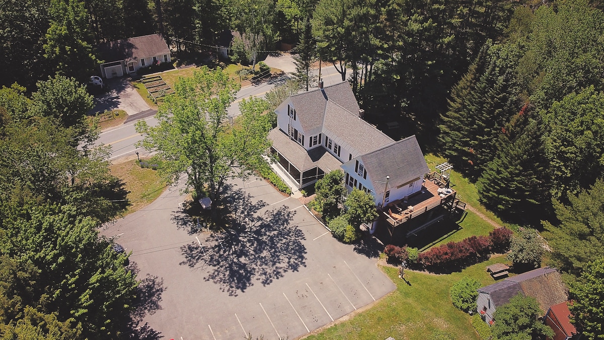 An aerial view reveals a spacious farmhouse-turned-hostel surrounded by trees and greenery. A large parking area is visible in the foreground, with multiple vehicles parked. The building features a deck on the upper level, offering an outdoor space for guests.