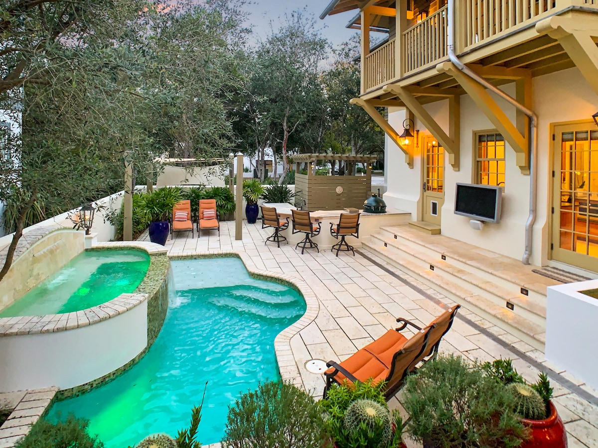 An inviting outdoor space features a turquoise pool alongside a spa, surrounded by well-maintained greenery and outdoor furniture. Several lounge chairs provide comfortable seating, and an outdoor kitchen area is visible, enhancing the relaxation options in this tranquil environment.