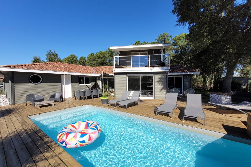 A contemporary villa features a private pool surrounded by a wooden deck. Sun loungers are arranged neatly along the edge, and a colorful inflatable float is present in the turquoise water. The house is designed with large windows and a balcony offering views of the serene garden.