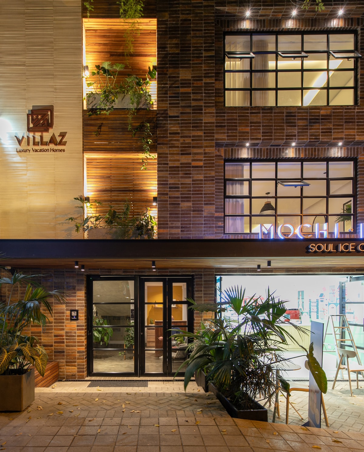 The entrance to Villaz Luxury Vacation Homes is illuminated with warm lighting, showcasing a modern façade of brick and wood elements. Large glass doors are framed by lush greenery and subtle plants, creating an inviting entry point to the property.
