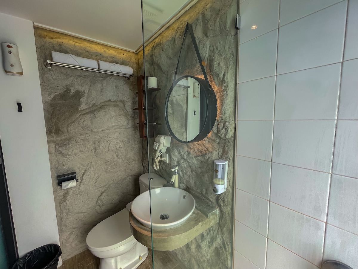 An eco-friendly bathroom features natural stone walls and modern fixtures. A circular mirror reflects soft lighting above a styled sink, and toiletries are neatly arranged. The area is equipped with a toilet and amenities, offering a clean and functional design.