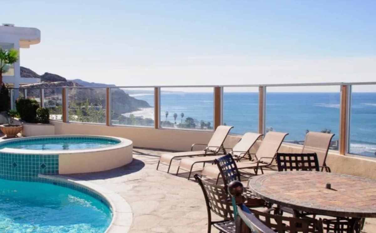 A private oceanfront patio features a hot tub and several lounge chairs, offering scenic views of the coastline. A round dining table stands nearby for outdoor meals, with glass railings providing an unobstructed vista of the ocean and surrounding landscape.