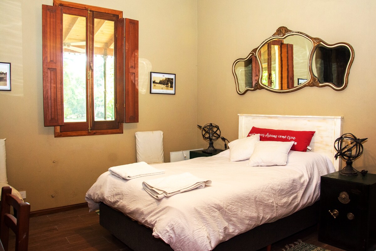 The guest room features a spacious bed with neutral-toned bedding and several pillows. Natural light enters through a wooden window framed by brown shutters. A decorative mirror hangs above the bed, and small tables are placed on each side. Framed images adorn the walls.
