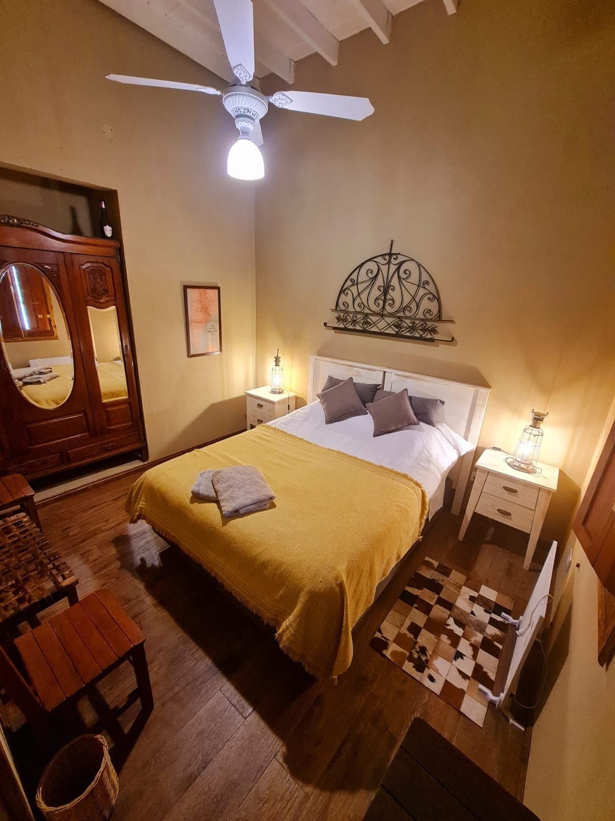 The cozy bedroom features a spacious bed covered in a yellow blanket, accompanied by decorative pillows. Two bedside tables with lamps flank the bed. A ceiling fan hangs above, and a mirror reflects the room's warm tones. A small wooden chair adds to the inviting atmosphere.