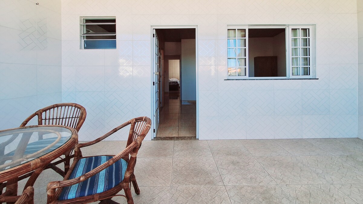 An outdoor space features a round glass table surrounded by four wicker chairs. A doorway leads into an interior area that appears well-lit. Large windows provide natural light, and the tiled floor offers a clean, smooth appearance.