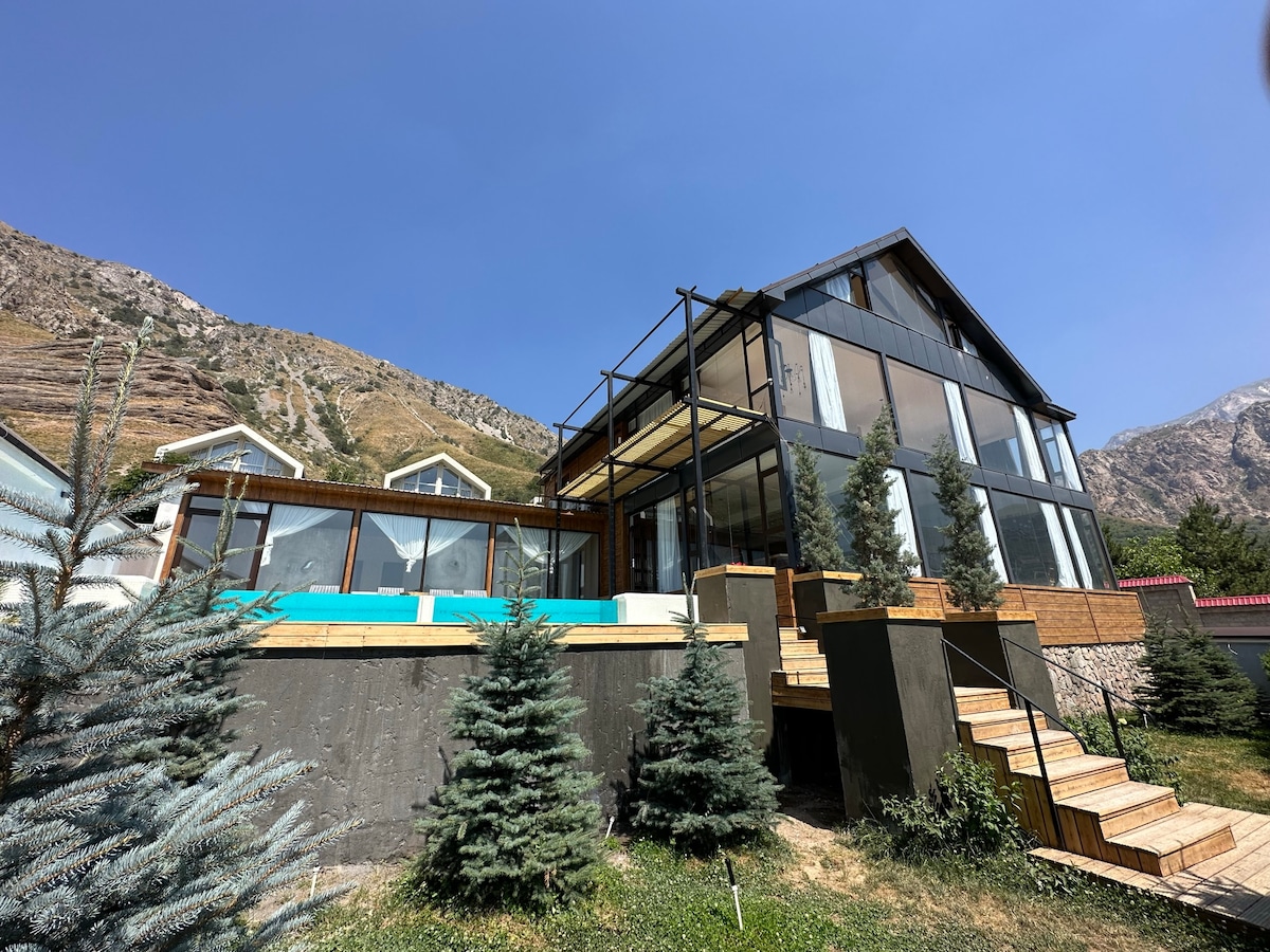 A modern villa features a large deck with a swimming pool, surrounded by manicured greenery and small trees. The structure showcases extensive glass windows, providing abundant natural light and views of the surrounding mountains.