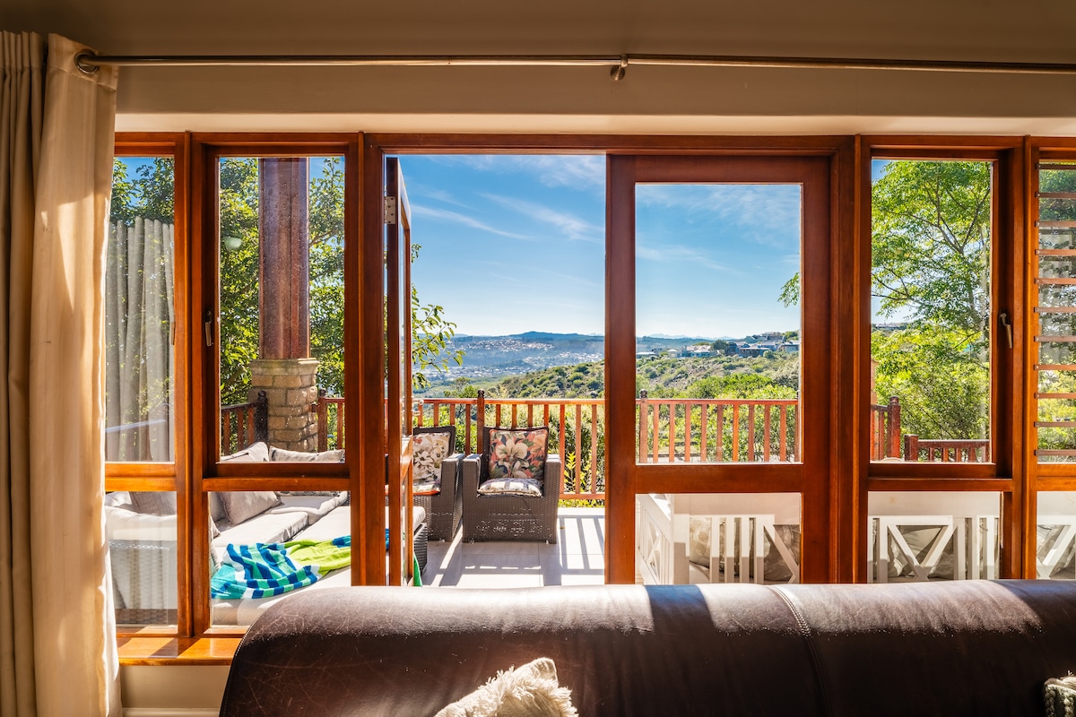 A spacious lounge area is visible, featuring a brown sofa that offers a view through large wooden windows. The outdoor deck is highlighted, showcasing seating arrangements against a backdrop of scenic greenery and distant hills under a clear blue sky.