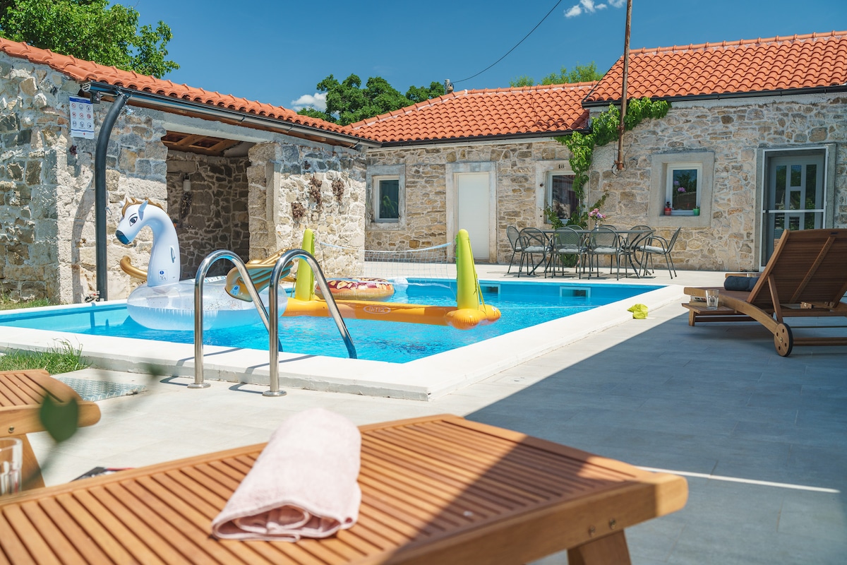 An outdoor area features a salt-water pool surrounded by a stone terrace, showcasing a sun lounger and a dining table with chairs. Colorful inflatable pool toys are present in the water, and a covered area is visible in the background, adding shade to the space.