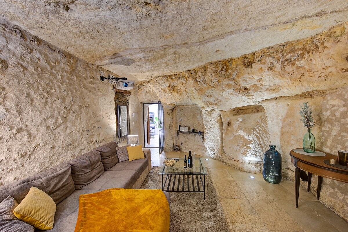 A unique troglodyte living area is presented, featuring a beige stone texture on the walls and ceiling. A sectional sofa surrounds a low coffee table, with a cozy blanket draped casually. Natural light enters through the doorway, highlighting decorative elements like a large vase and a small table.