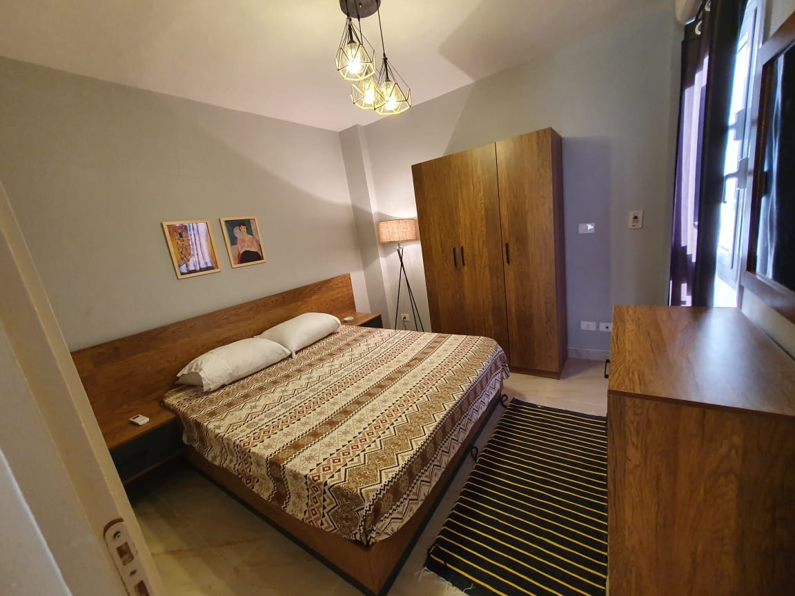 A bright bedroom features a spacious bed with a patterned blanket. A wooden wardrobe and dresser provide ample storage. A lamp stands beside the bed, and two framed artworks adorn the wall. Natural light filters in through a window with sheer curtains.