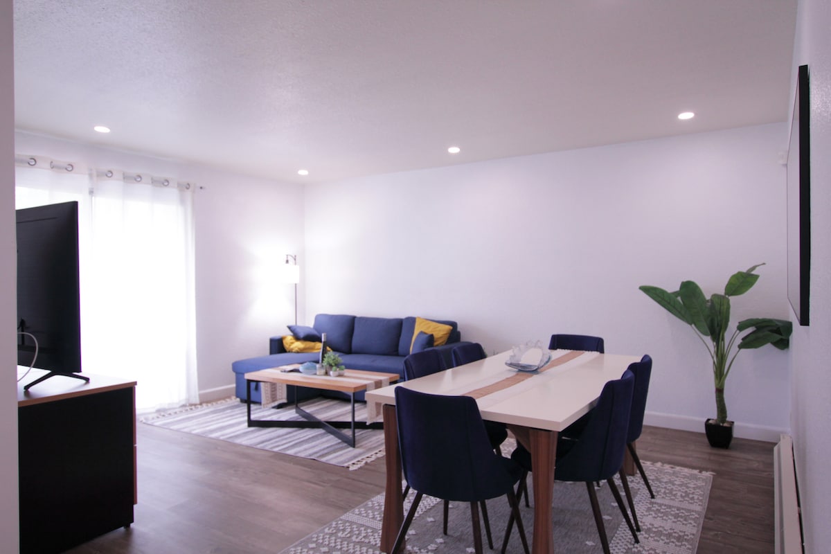 A modern living and dining area features a navy sofa adorned with yellow pillows. A wooden dining table is surrounded by six dark blue chairs, providing ample seating. A large television stands on a light-colored media console, and natural light enters through sheer curtains.