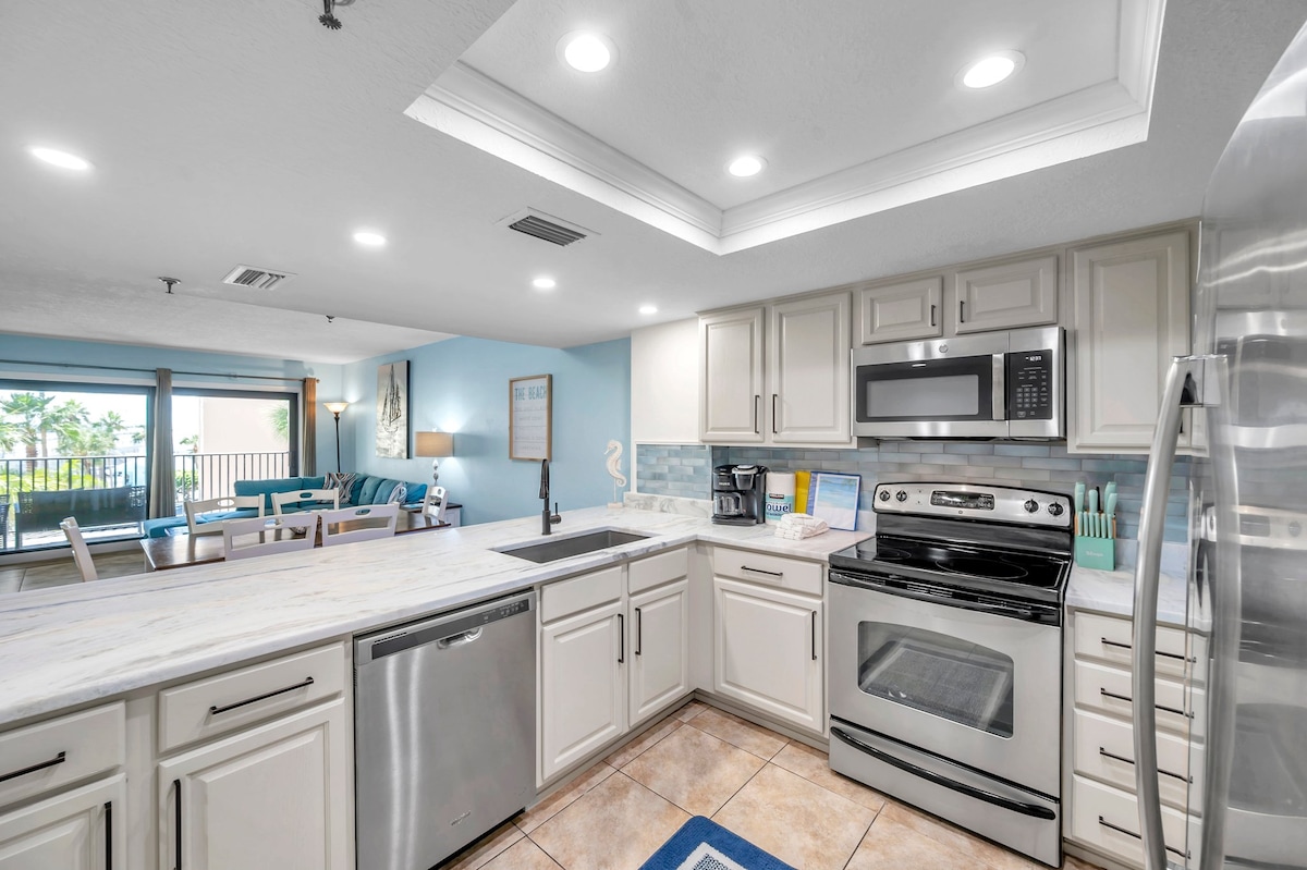 A modern kitchen arrangement features sleek stainless steel appliances, including a stove, microwave, and dishwasher. Light-colored cabinetry is paired with a large stone countertop. The open space is well-lit with recessed lighting, and views of the balcony area are visible through the nearby sliding glass doors.