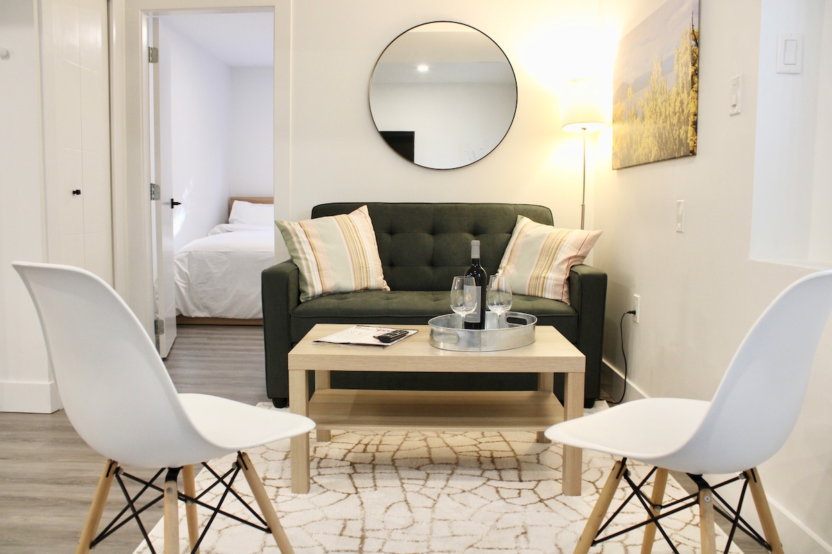 A comfortable living area features a green sofa with decorative pillows, positioned across from two white chairs. A small wooden table holds glasses and a bottle, while a round mirror reflects light. In the background, the entrance to a bedroom is visible.