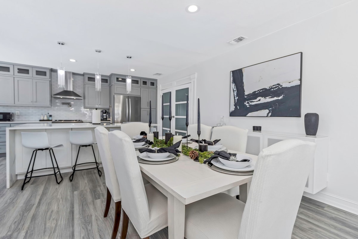 A spacious dining area is furnished with a light-colored table surrounded by upholstered chairs. A set of black candles stands at the center of the table, complemented by greenery. Modern bar stools line the kitchen counter, and natural light illuminates the room.