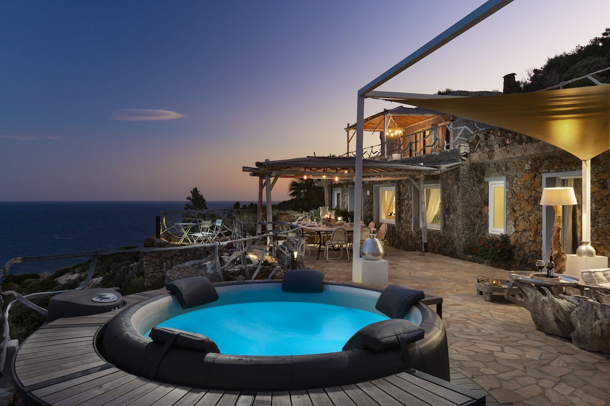 A spacious outdoor terrace is highlighted by a bubbling hot tub, surrounded by natural stone walls. Relaxing seating areas and a dining table are present, offering stunning ocean views at dusk. Soft lighting enhances the ambiance, while the sea stretches in the background.