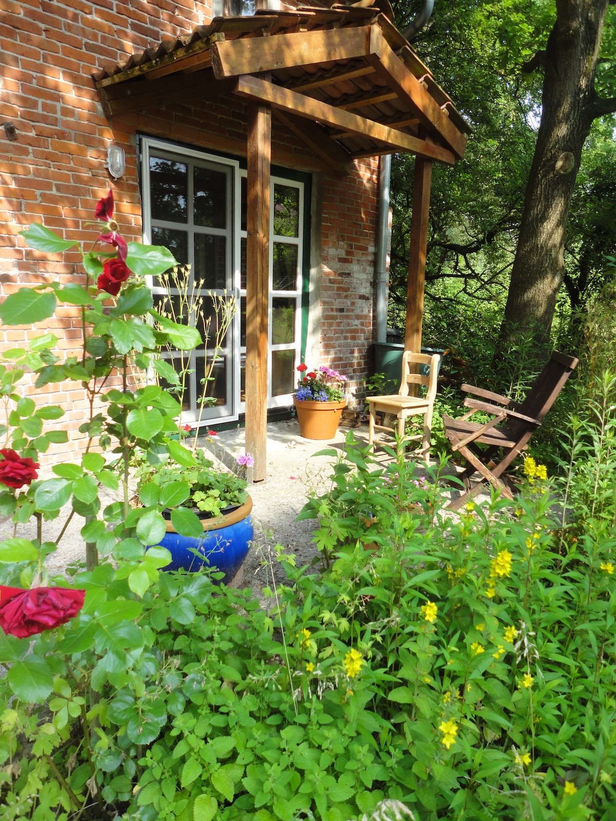 A welcoming entrance is framed by a wooden overhang, featuring a small table and a chair beside potted flowers. Lush greenery and colorful blooms surround the space, creating a serene outdoor environment. The entrance showcases large glass doors that provide a glimpse into the interior.