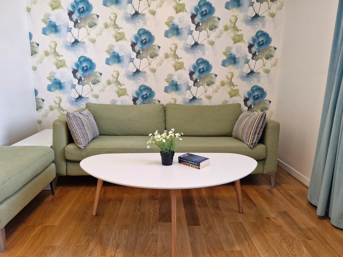 A green sofa is positioned against a floral wallpaper featuring blue flowers and soft colors. A round white coffee table is placed in front, adorned with a small potted plant and a couple of books. Natural light illuminates the wooden floor.